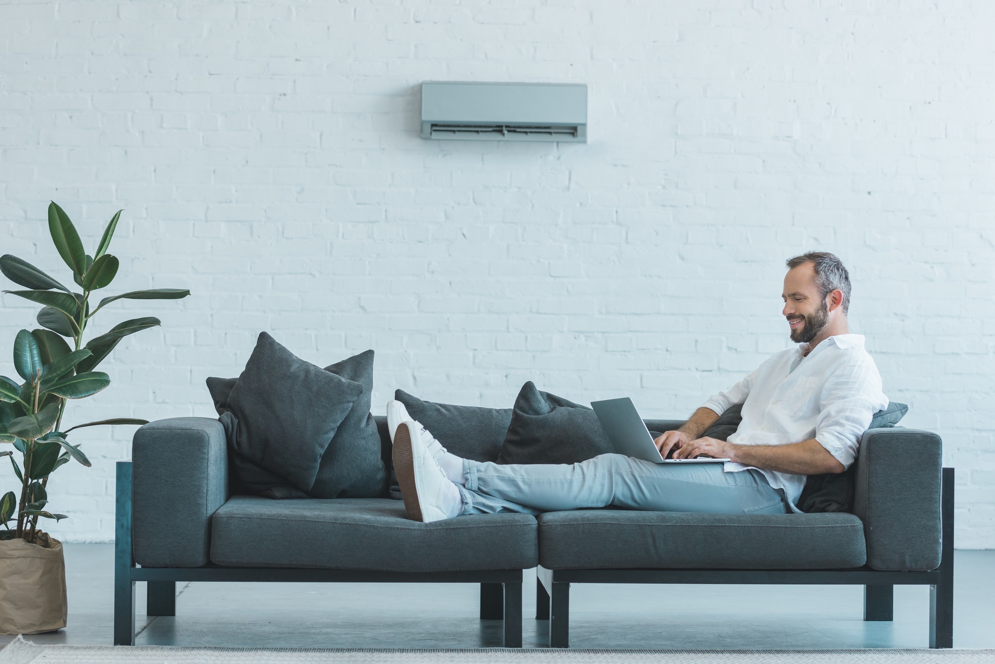 Man working on a laptop on sofa with air conditioner on the wall of home.